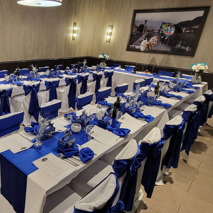 Long dining tables set with white tablecloths, blue runners, and blue chair sashes for an event.