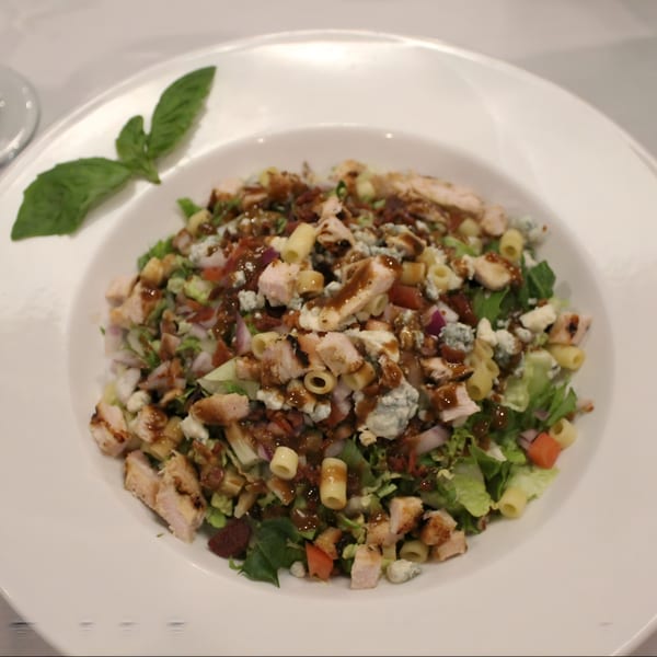 A white bowl filled with a chopped salad, chicken, pasta, and blue cheese, drizzled with dressing.